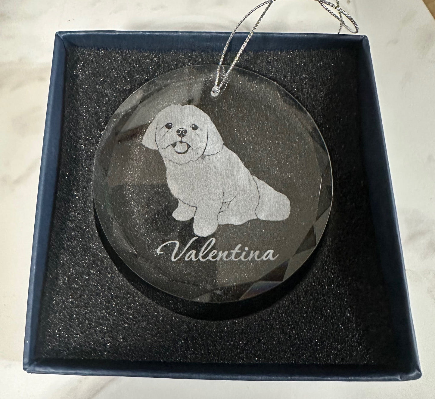 Decorative glass ornament with engraved dog design and 'Valentina' text, in a box on a marble surface.